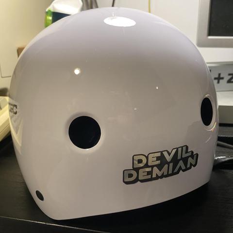 A white skating protective helmet seen from the back. Near the bottom lip, a “Devil Demian” sticker is applied.