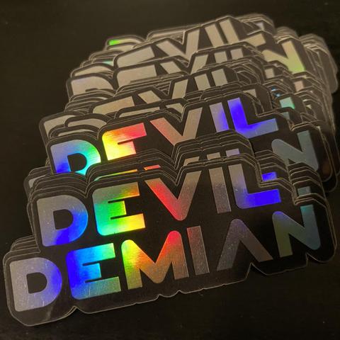 A stack of stickers. It’s shape is formed by the words “Devil Demian”, one of top of the other. The colors of the rainbow refract from its metallic surface.