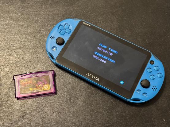 A Picture of Shantae Advance with a PS Vita next to it. The vita is showing the game's completion screen with a time of 6h 30m with a completion of 98/100