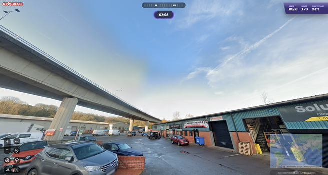 Screenshot aus GeoGuessr, man sieht links eine Straße auf Betonstelzen und rechts ein einstöckiges Gewerbegebäude, Wände unten aus Ziegeln und oben aus Wellblech, etwas im Hintergrund ist zu lesen "Cardiff Remap - Tuning Specialists". Ansonsten hauptsächlich eine große Asphaltfläche, auf der mehrere Autos herumstehen.