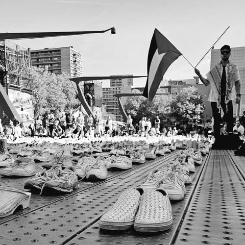 Een zwart-witfoto van een plein waar honderden paar schoenen netjes in rijen zijn geplaatst, symbool voor de vermoorde Palestijnse zorgverleners in Gaza. Op de achtergrond staan mensen bijeen, en rechts hou ik een Palestijnse vlag omhoog, gekleed in een keffiyeh. De scène vindt plaats in een stedelijke omgeving met hoge gebouwen en bomen.