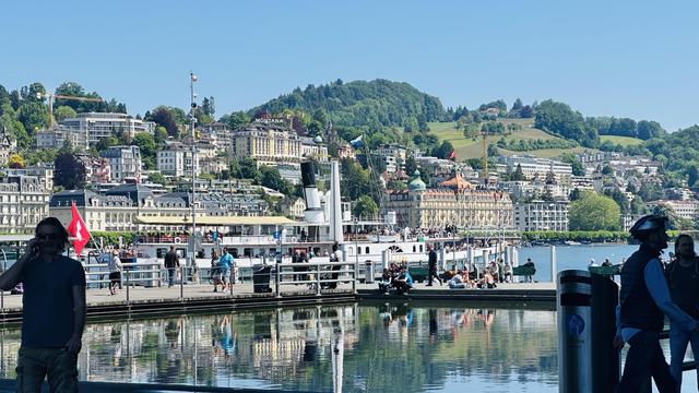 Ein Schiff verlässt gerade den Hafen von Luzern, dahinter sieht man einen Teil der Stadt mit teuren Hotels und Villen.
Darüber wolkenloser, blauer Himmel.
