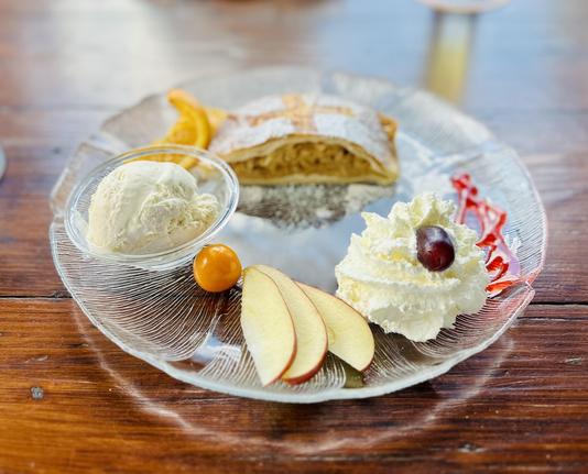 Apfelstrudel mit Vanilleglacé auf Glasteller. Reichhaltig dekoriert mit Orangenscheibe, Apfelstückchen, Schlagsahne mit Traube und Kumquat.
