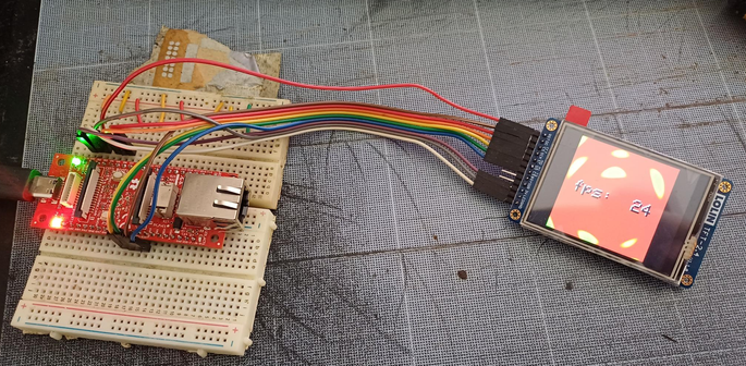 An esp32p4 development kit from Olimex is sitting between two breadboard (because it wouldn't fit on just one). Flying wires are connected one one side to the breadboard, and on the other side to a small display panel, where a plasma effect along with the text "FPS: 24" can be seen.
