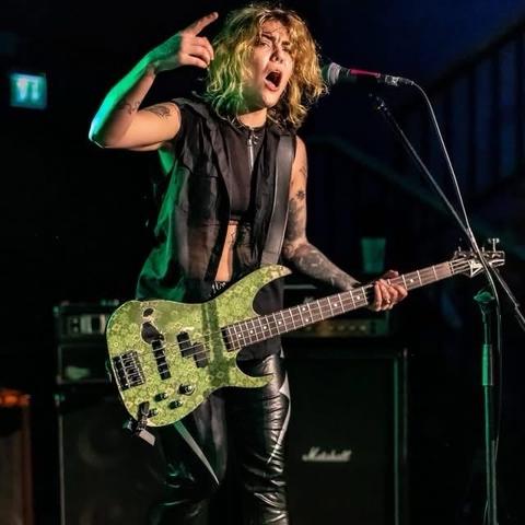 Eine Musikerin mit welligen, blonden Haaren tritt auf der Bühne auf und hält leidenschaftlich eine grüne Bass-Gitarre in der Hand. Sie trägt ein schwarzes ärmelloses Oberteil und eine Lederhose, umgeben von Verstärkern an einem dunkel beleuchteten Ort.