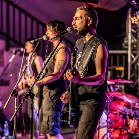 Eine Live-Musikaufführung mit drei Musiker:innen, die singen und E-Gitarren spielen. Sie stehen auf der Bühne mit Mikrofonen und einem im Hintergrund sichtbaren Schlagzeug. Die Beleuchtung ist farbenfroh und schafft eine lebendige Atmosphäre.