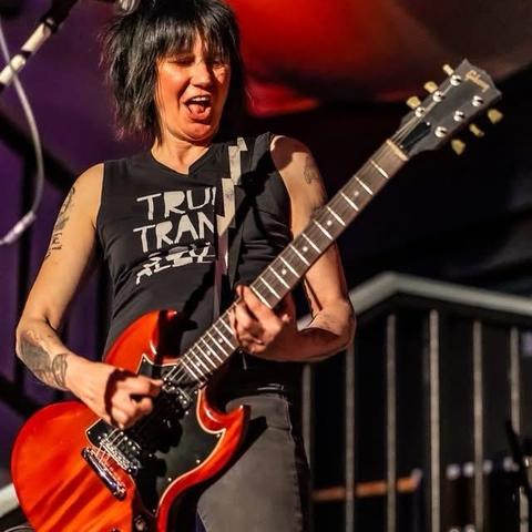 Eine Gitarristin tritt auf der Bühne auf, trägt ein ärmelloses schwarzes Hemd, spielt eine E-Gitarre mit einem ausdrucksstarken Gesichtsausdruck. Der Hintergrund zeigt Bühnenbeleuchtung und Ausrüstung.