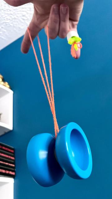 A blue Luftverk Plastic Peak yo-yo, set up for 5A (freehand) play, spinning after a successful suicide catch. A teal wall overtakes most of the background with only a sliver of a white book shelf visible to the left of the frame. 