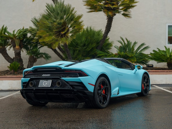 a photo of the vehicle seen from behind in a 3 quarter view, the convertible top down, it is in  a parking lot, shubs growing against the building in the background 