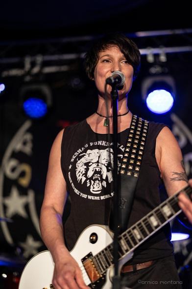 A woman with short hair performs on stage, holding a microphone and an electric guitar. She wears a sleeveless black shirt with a graphic design and a studded strap on her guitar. Colorful stage lights are visible in the background.