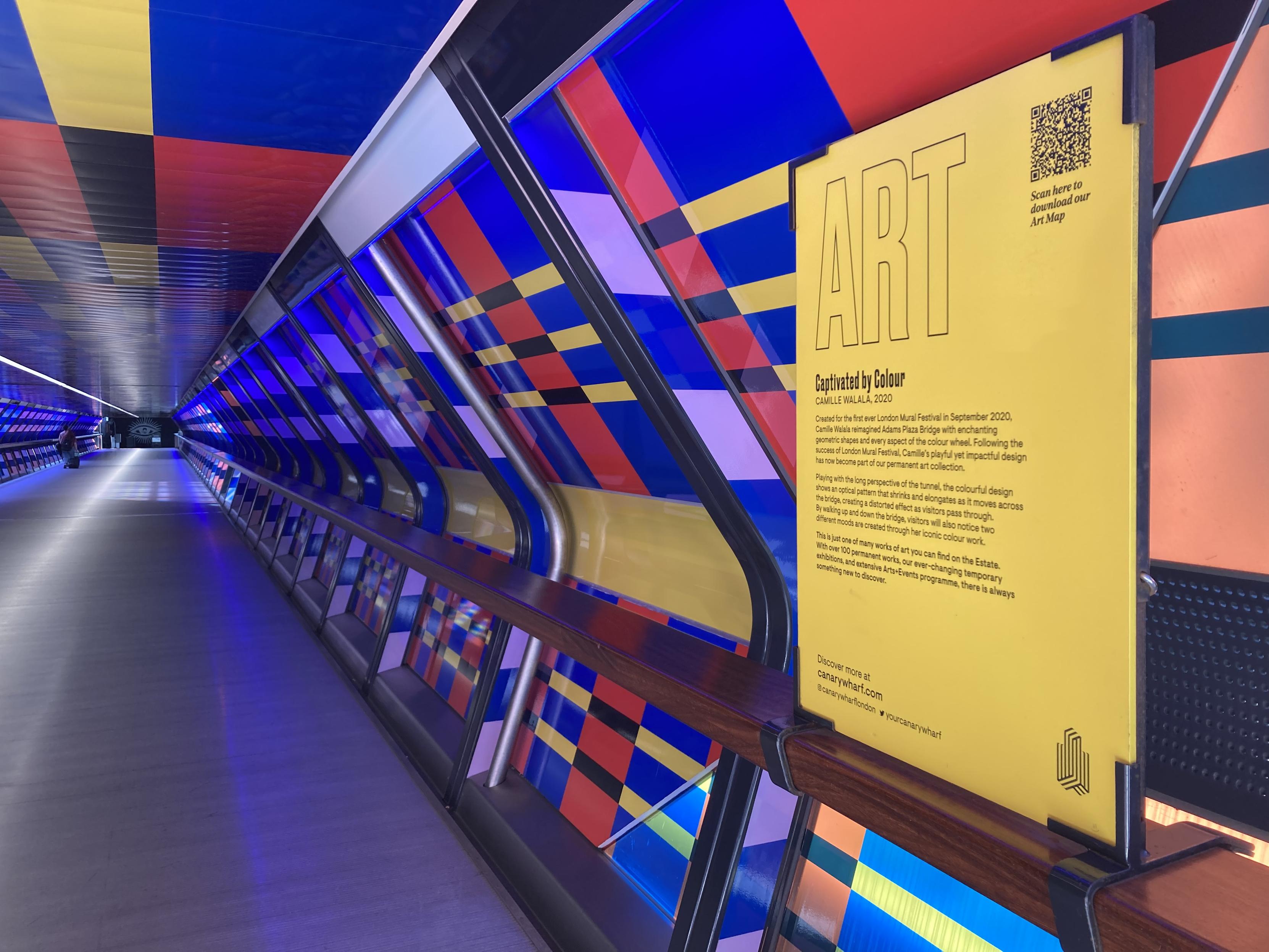 the colourful bridge over Adam's Place in Canary wharf, with a geometric pattern on the walls and ceiling. on the right-hand side, the photo includes the artwork description:

ART
Captivated by Colour
CAMILLE WALALA, 2020
Created for the first ever London Mural Festival in September 2020, Camille Walala reimagined Adams Plaza Bridge with enchanting geometric shapes and every aspect of the colour wheel. Following the success of London Mural Festival, Camille's playful yet impactful design has now become part of our permanent art collection.
Playing with the long perspective of the tunnel, the colourful design shows an optical pattern that shrinks and elongates as it moves across the bridge, creating a distorted effect as visitors pass through.
By waLking up and down the bridge, visitors will also notice two different moods are created through her iconic colour work.
This is just one of many works of art you can find on the Estate.
With over 100 permanent works, our ever-changing temporary exhibitions, and extensive Arts+Events programme, there is always something new to discover.
Scan here to download our
Art Map
Discover more at canarywharf.com