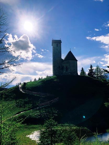 Una chiestta su di una collina in montagna con il sole alto in cielo. Sotto si trova un piccolo laghetto