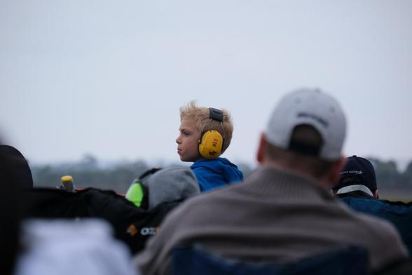 Photo of a young boy in a blue hoodie wearing large, yellow hearing protection earmuffs while watching the flying display at an airshow. 