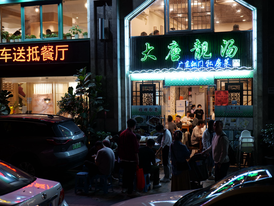 Neon signs light up the front of two restaurants in Shenzhen, China. People mill around outside of the restaurant on the right waiting to get in