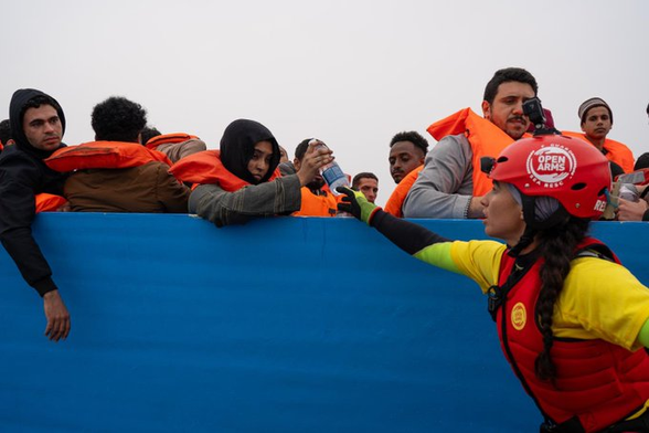 Menschen sitzen dichtgedrängt in einem blauen Holzboot. Sie sind mit Rettungswesten versorgt. Eine Seenotretterin reicht ihnen Wasser.
