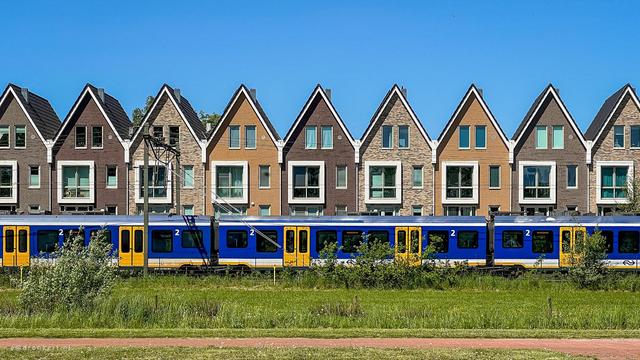A row of 9 terraced houses with pointy roofs how a child would draw a house. They look new and identical except for different brick colours. In front of them a train drives by, it is dark blue with white stripes up top and below, with yellow doors. Closer still, some grass and red asphalt. Clear blue sky. The symmetry of the houses is slightly broken because the row was not quite perpendicular to my view.