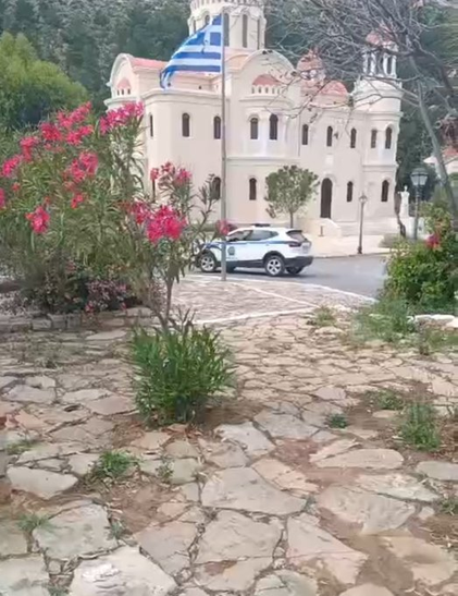 Aus einer Grünanlage heraus aufgenommen: Eine größere Kirche mit einem Fahnenmast davor, an dem die griechische Nationalfahne weht. Daneben steht ein PKW der Hellenic Police. Hinter der Kirche steigt das Gelände steil an und ist dicht bewachsen.