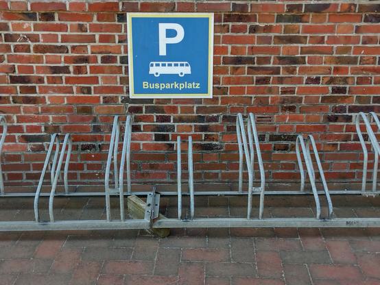 Das Bild zeigt eine Parkanlage für Busse an einer roten Backsteinwand. Im Vordergrund befindet sich ein Metallgerüst, das für die Parken von Fahrrädern vorgesehen ist, obwohl es leer ist. Über dem Gerüst ist ein blaues Schild mit einem weißen "P" und einem Bus-Symbol, das "Busparkplatz" in gelber Schrift darunter besagt. Die Wand besteht aus roten Backsteinen, die in verschiedenen Farbtönen und Größen angeordnet sind. Der Boden besteht aus roten Ziegeln, die in einem geometrischen Muster angeordnet sind. Das Schild und das Gerüst sind in einem guten Zustand, und die Umgebung wirkt sauber und gut gepflegt.