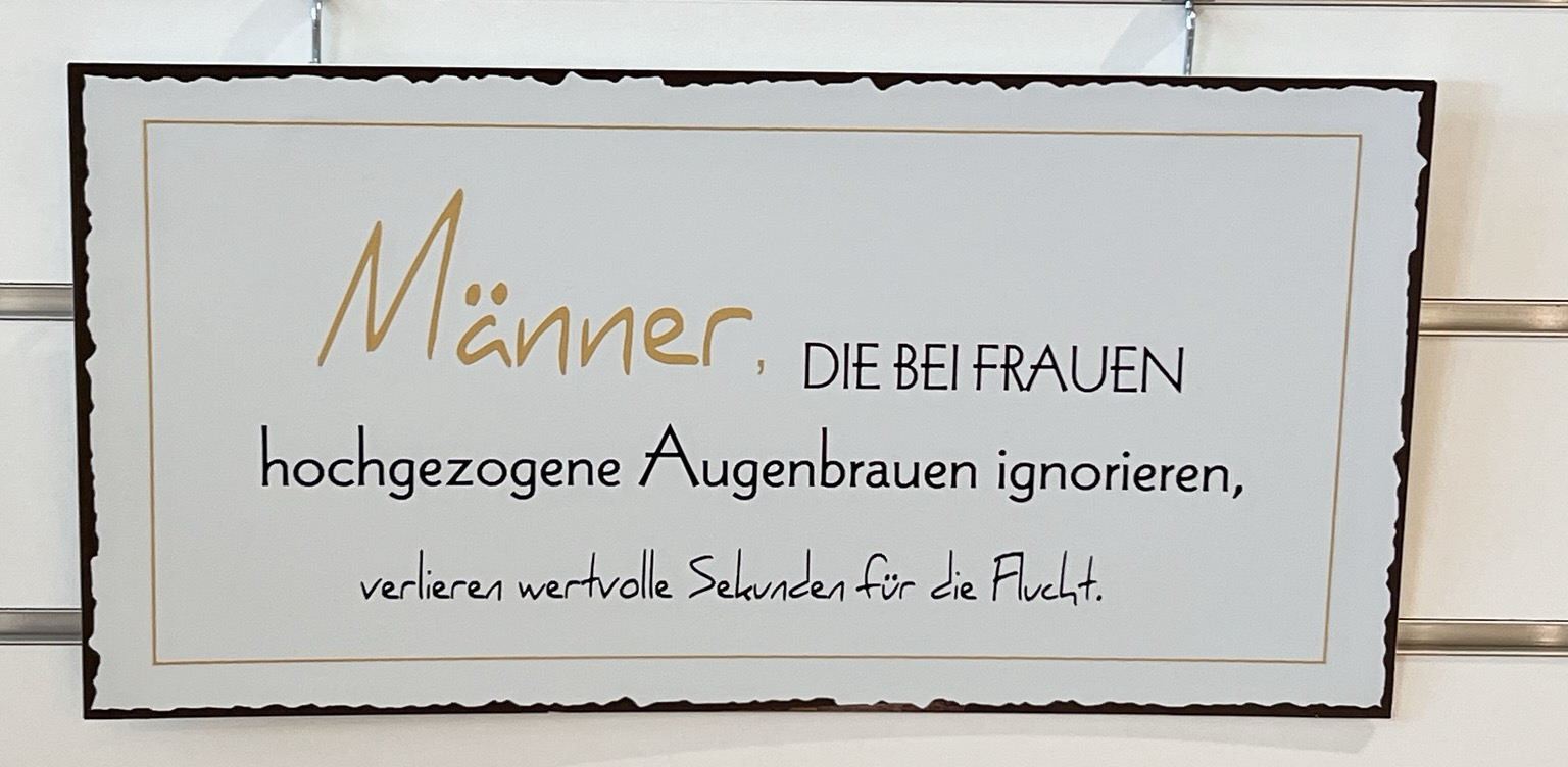 Farbfotografie im Querformat zeigt ein weißes Schild mit gelber und schwarzer Srift, dass an 2 Drähten an einer weißen Wand befestigt ist.  In gelb ist das Wort "Männer" geschrieben, der restliche Text ist in schwarzer Schrift. Auf dem Schild steht der folgende Text: "Männer, DIE BEI FRAUEN hochgezogene Augenbrauen ignorieren, verlieren wertvolle Sekunden für die Flucht. 