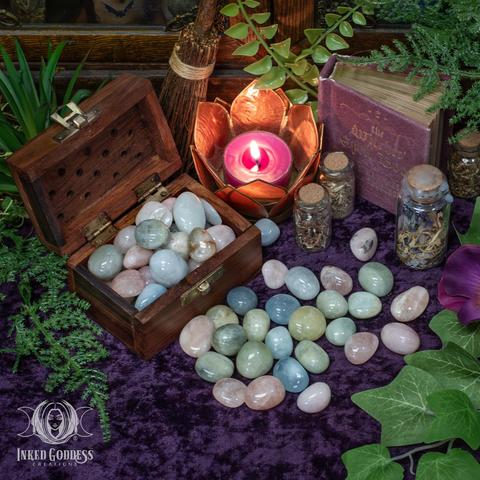 Several specimens of the "Morganite Tumbled Gemstone" product are arranged on a dark velvet-covered surface. Several more specimens fill a wooden box with a hinged lid. They are in pastel shades of pink, green, and blue and range from translucent to opaque. They are round, tumbled smooth, and polished. Presented by Inked Goddess Creations.