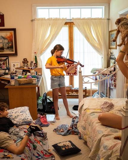 Photo of a child playing viola in a messy bedroom. Another child is lying nearby on the ground, and a person out of frame is sitting on a bed holding an animal over their head.