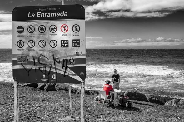 Schwarzweissfotografie mit Selektivfarbe Rot eines grossen Schilds mit vielen Piktogrammen. Eines davon ist "No Lifeguard", am Ufer sitzt aber eine Person an einem Tisch mit einem roten Shirt mit der Aufschrift "Lifeguard". 