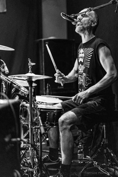 A drummer in a sleeveless shirt plays energetically on stage, with a microphone in front of him. He appears focused and expressive, surrounded by drums and equipment, in a black and white setting.