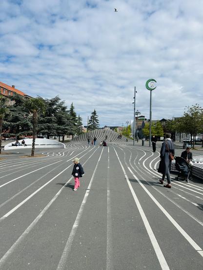 A pedestrian area featuring wavy, striped walkways. People are walking, including a child in pink pants. Lush trees and buildings are visible in the background under a cloudy sky.
