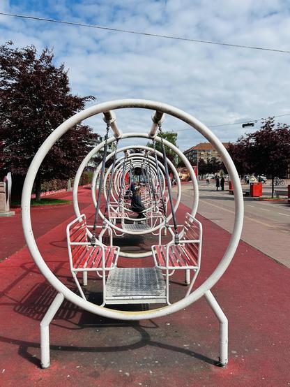 A row of circular swings is shown in a playground setting, with a person sitting on one swing. The ground is covered in red material, and there are trees and buildings in the background under a cloudy sky.