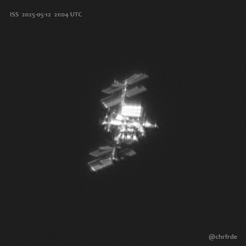 White ISS against black background. The central axis with the main modules shows somewhat splotchy and partially overexposed structures. Above and below four large solar panels appear darker but still well illuminated in this perspective. Direction of movement is toward the left.