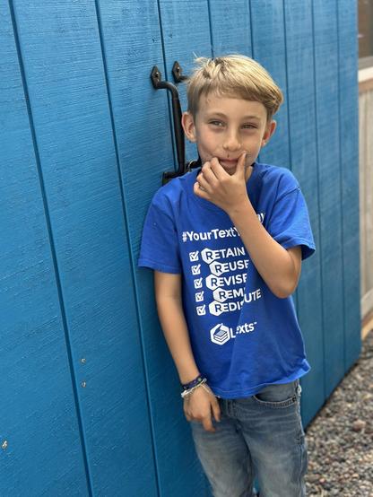 Young boy with blonde hair standing in front of a bright blue shed door while wearing a LibreTexts shirt and blue jeans, smiling as he holds his hand near his mouth 