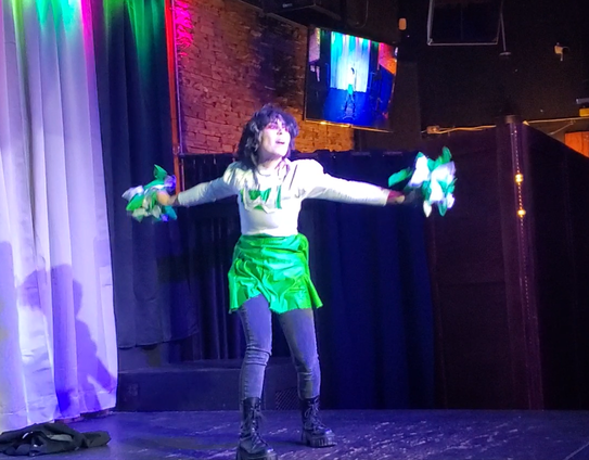 Austin Tacious onstage at the Junction pub, and dressed in Gerard Way's white and green cheerleader dress. His hair is spray-painted black and flat-ironed, and he's wearing smudged red eyeliner, so he looks a bit dead. He's gesturing wildly with cheerleading pompoms in outstretched arms.