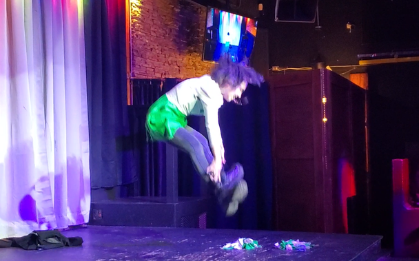 A very blurry photo of Austin Tacious, dressed as Gerard Way as a cheerleader, jumping into the air and touching his toes.