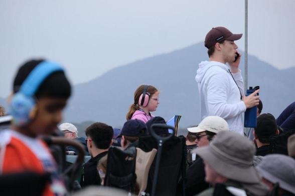 Photo of a young girl in a pink hoodie wearing large, pink hearing protection earmuffs while watching the flying display at an airshow. 