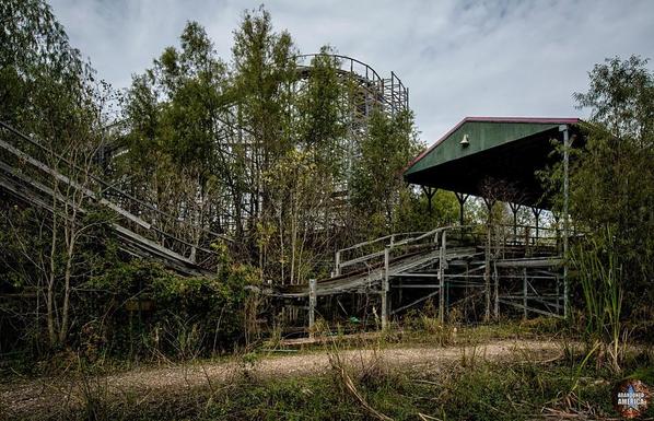 A wooden boarding pavilion is on the right with dilapidated tracks exiting it to the left. A high loop is visible behind them. They are overgrown by trees and weeds.