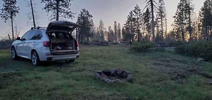 A white SUV next to a camp fire, parked on grass among trees.