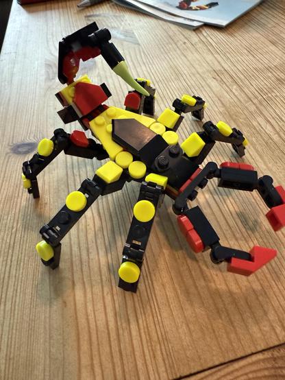 A colorful scorpion model made from interlocking plastic pieces, featuring yellow, black, and red components. The scorpion is posed with its pincers raised on a wooden surface.