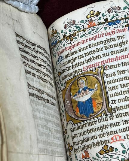 Een pagina uit een middeleeuws manuscript met een rijk versierd initiaal waarin een man een doek met drie baby's vasthoudt. De rand van de pagina is versierd met gekleurde en gouden bloemen. 