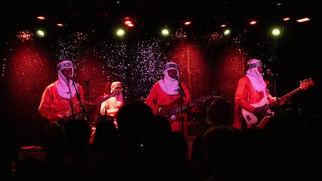 Four members of a rock band on stage, red-lit by stage lighting. They wear red kaftans and white headdresses. Three have electric guitars, while the fourth plays drums.