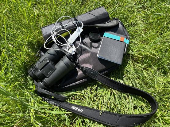 Foto einer Fahrradtasche für den Fahrradrahmen auf der Equipment liegt. Es ist ein Fernglas, Kopfhörer mit integrierten Mikrofonen, ein kleiner Fahrradcomputer  und ein Notizbuch mit türkisem Band und Stift zu sehen. 