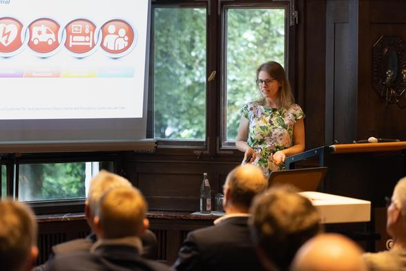 Dr. Camilla Metelmann ist Ahnefeld-Stiftungsprofessorin für Notfallmedizin. Auf dem Bild zu sehen ist eine junge Frau mit langen Haaren und einem geblümten Kleid, die in einem rustikalen Festsaal einen Vortrag hält. Von hinten zu sehen sind einige Männer aus der Zuhörerschaft. 