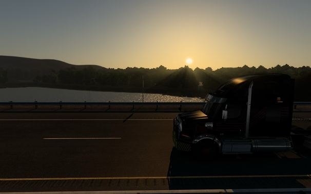 A black semi-truck is parked on the side of a highway during sunset. The sun is low in the sky, casting a warm glow over the landscape. In the background, there is a body of water surrounded by trees and hills. The scene is tranquil and picturesque, highlighting the beauty of nature and the peacefulness of the evening.