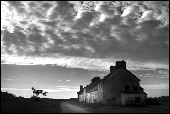 La photo est en noir et blanc. On voit un bâtiment en ruine éclairé par la gauche. Le reste est dans l’ombre, tout comme le paysage dans lequel on se trouve. Seule la route étroite qui passe devant le bâtiment est légèrement éclairée elle aussi. Le ciel occupe presque toute l’image. La lumière particulière vient de la présence d’un gros nuage qui occupe le moitié supérieur de l’image, mais qu’en même temps un rayon de soleil éclaire la bâtisse. 