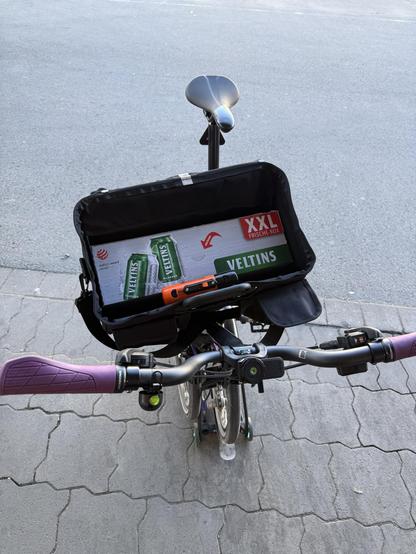 A bicycle is parked on a paved surface, featuring a saddle and a handlebar with purple grips. A black storage bag is attached to the back, holding a box of Veltins beer and an orange tool. The area appears to be a street