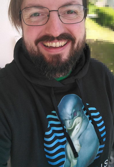 A middle aged German streamer making bad choices, smiling while he wears a black hoodie with a blue half naked Dolphin person on it from the game Stellaris.