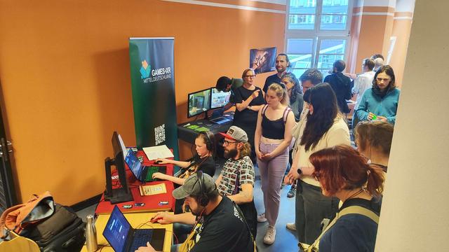 People standing in the corridor of a university, rows of tables with games where people casually play video games.