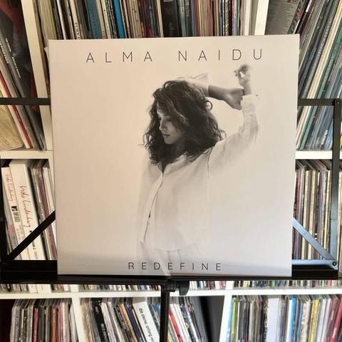 Ein Albumcover, das auf einem Ständer ausgestellt ist, zeigt ein Schwarz-Weiß-Bild einer Frau in einem weißen Hemd, mit dem Titel "REDEFINE" und dem Namen der Künstlerin "ALMA NAIDU", prominent dargestellt. Der Hintergrund zeigt eine Sammlung von Schallplatten.
