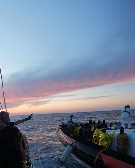 Eine Rettungsaktion auf See bei Sonnenuntergang oder Sonnenaufgang. Vom Deck eines größeren Schiffes aus sieht man den Bug einer Guardia Costiera CP, ein Schlauchboot mit festem Rumpf, in dem sich zahlreiche Personen befinden, einige davon in goldene Rettungsdecken gehüllt. Von hinten zu sehen: Eine Person in weißer Schutzkleidung und Rettungsweste hantiert an der Reling der CP. Eine Person im Vordergrund streckt einen Arm zu einem Abschiedsgruß aus. Der Himmel ist in Rosa- und Blautönen gefärbt, das Meer ist relativ ruhig.