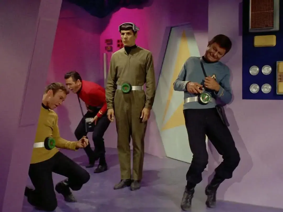 This is a scene from the original Star Trek television series, known for its colorful, futuristic set design and dramatic storytelling. The setting features a retro-style spaceship interior with bright pink and purple walls and angular, geometric architecture—characteristic of 1960s sci-fi.

Four characters are in the scene:

    Captain Kirk (in a mustard-yellow uniform) is kneeling on the floor, clutching at his chest in apparent pain or dramatic reaction.

    Scotty (in red) is slightly bent over, also appearing to be in discomfort or reacting to something forceful.

    Spock (in a solid olive-green outfit and a headband-like device) stands calmly and rigidly in the center, seemingly unaffected and observing the others.

    Dr. McCoy (in a light blue uniform) leans against the wall to the right, also clutching at his chest or belt area, possibly in pain or strain, grimacing.

All of them are wearing silver belts with green circular devices that appear to be causing them some kind of reaction—perhaps as part of the plot's sci-fi technology or alien influence. The scene likely involves some external force affecting the crew, with Spock remaining stoic and composed, in contrast to the visibly struggling humans.

The overall tone is dramatic, yet the vibrant colors and exaggerated expressions give it a slightly surreal or campy feel, typical of early Star Trek episodes.