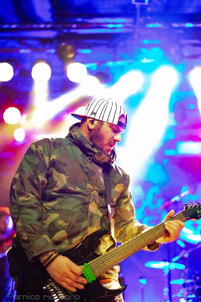 A musician in a camouflage jacket and a baseball cap plays an electric guitar on stage, illuminated by colorful stage lights. The image captures an energetic performance atmosphere.